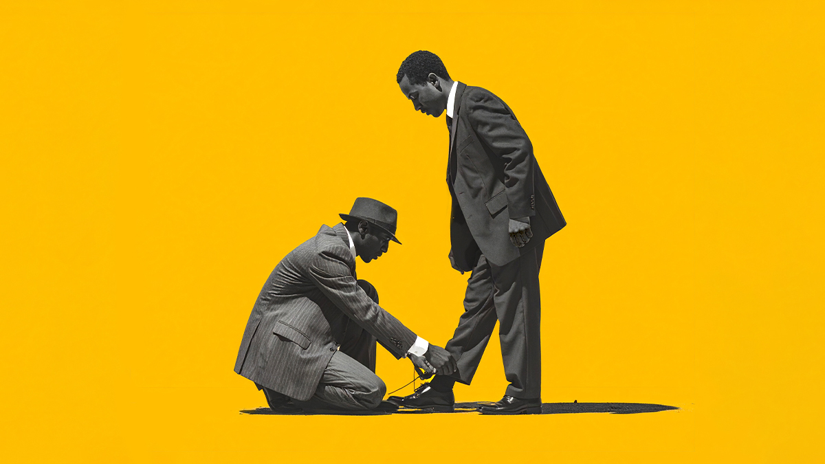 A photo 2 people in suits one tying the others shoes against a yellow background.