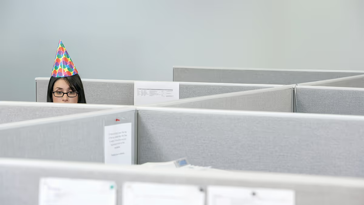 Photo of a person standing in a cubicle with a party hat on.