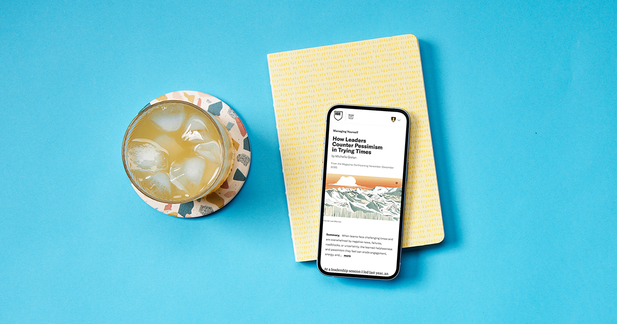 Overhead view of a smartphone showing a Harvard Business Review article on a bright blue desktop next to an iced drink and a notebook.