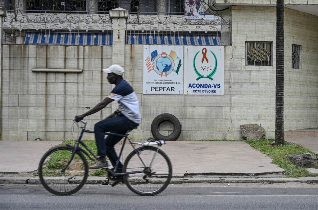 A man on a bicycle rides past a building wall in Abidjan, Côte d'Ivoire, which has signs for PEPFAR (the U.S. President's Emergency Plan for AIDS Relief) and ACONDA-VS, a local Ivorian AIDS relief organization.