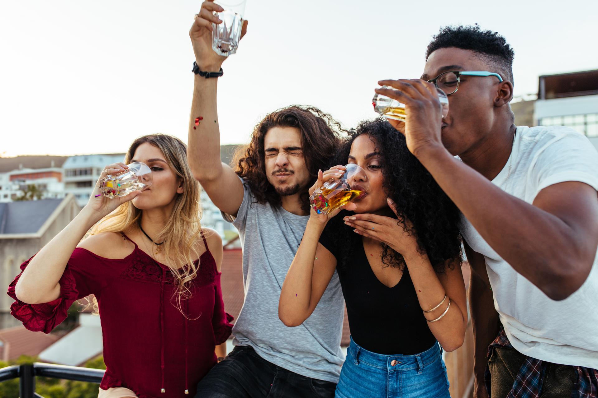 A white woman, Latino man, Black woman, and Black man drink beer as if chugging it on a sunny rooftop.