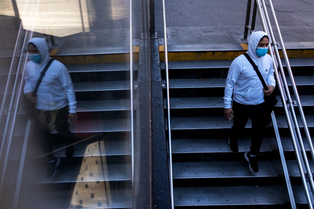 A person wearing a mask walking down a flight of stairs to a subway stop, reflected in a mirror so it is a double image.