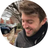 Miles, a white man with a short brown beard and a shock of brown hair smiles broadly as he eats a giant cookie.