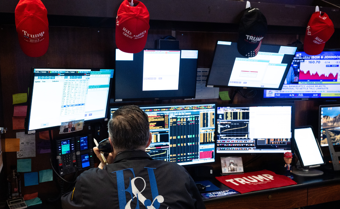 Rearview of a stock trader looking at his mutiple screens with MAGA hats and Trump paraphernalia surrounding him