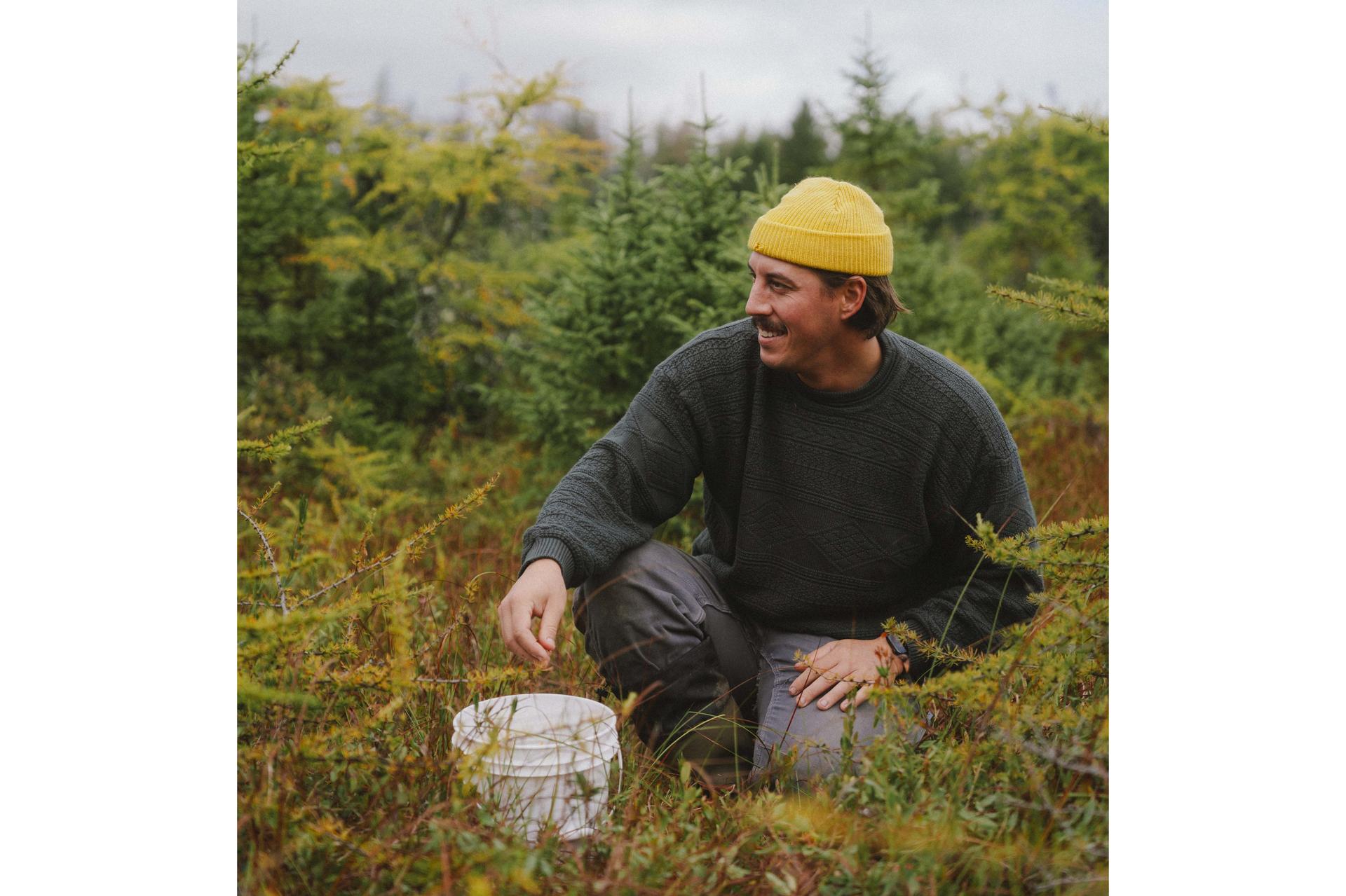 A man in a yellow beanie, dark grey sweatshirt and grey pants, sitting in a lush field with a white bucket