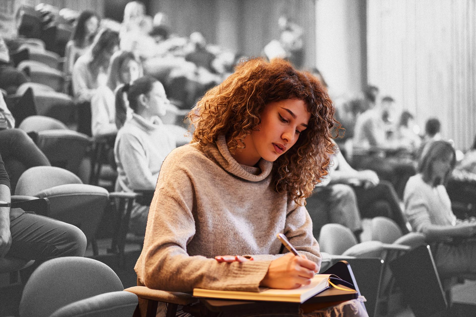 A woman with curly hair, wearing a turtleneck and writing in a notebook in a classroom filled with students 