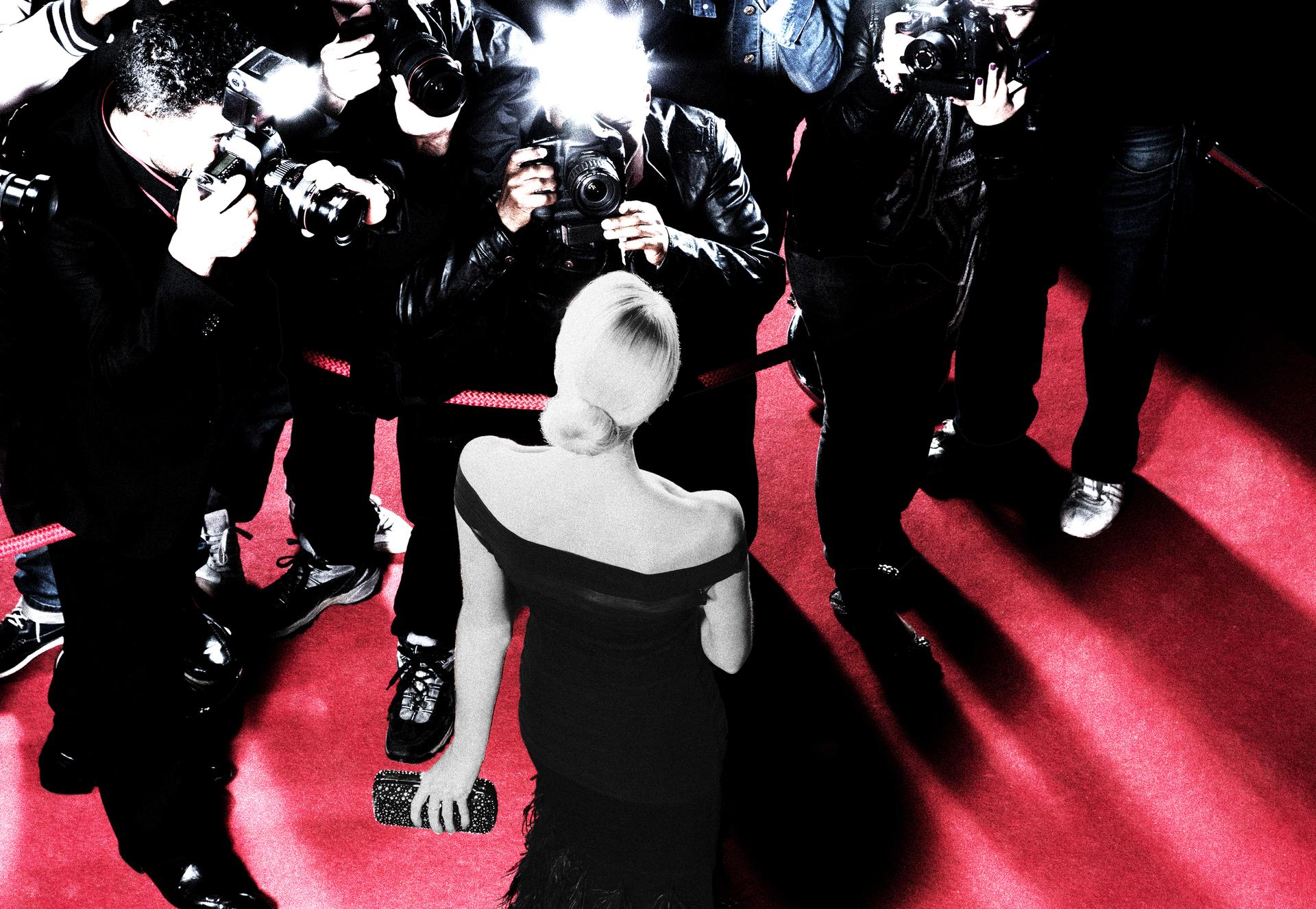 A blonde woman in a black dress standing on a red carpet, being photographed by paparazzi