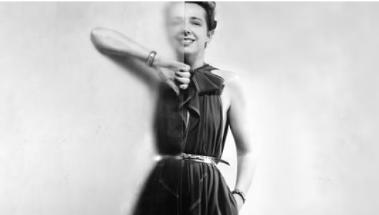 A black-and-white portrait of Claire McCardell, standing partially behind a frosted glass panel in a black dress, her left hand tucked into a side pocket.