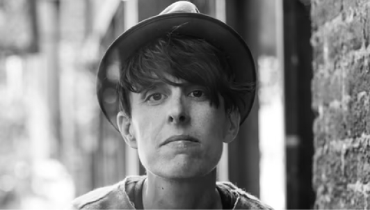 A black-and-white photo of a person with dark hair wearing a narrow hat and a thin sweater beside a brick wall
