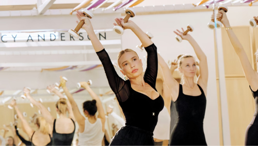 photo of reflection in mirror of Anderson leading a fitness class with both arms raised above head and hands holding weights in well-lit studio