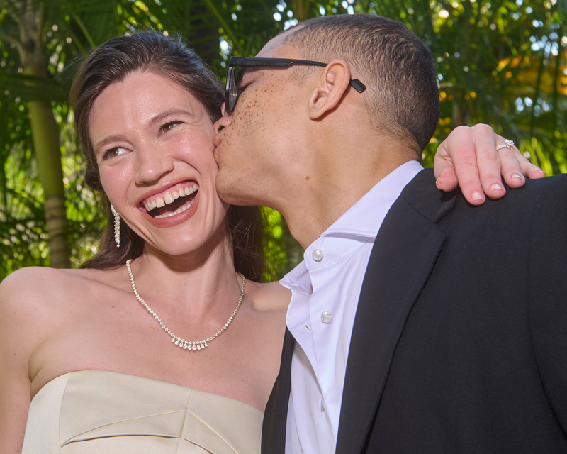 A joyful couple laughing and sharing a kiss, elegantly showcasing stunning diamond necklaces and sparkling earrings.