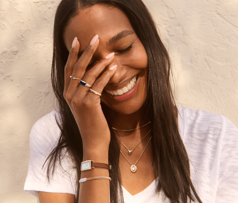 A woman laughs, resting her face in her hand to reveal stunning rings, a watch, bracelet, and several necklaces