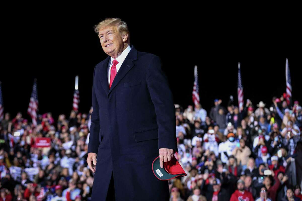Donald Trump stands in the foreground in a long black coat with a red tie, holding what appears to be a red MAGA hat. In the background is a crowd of people, a row of American flags and the dark black of a night sky. 