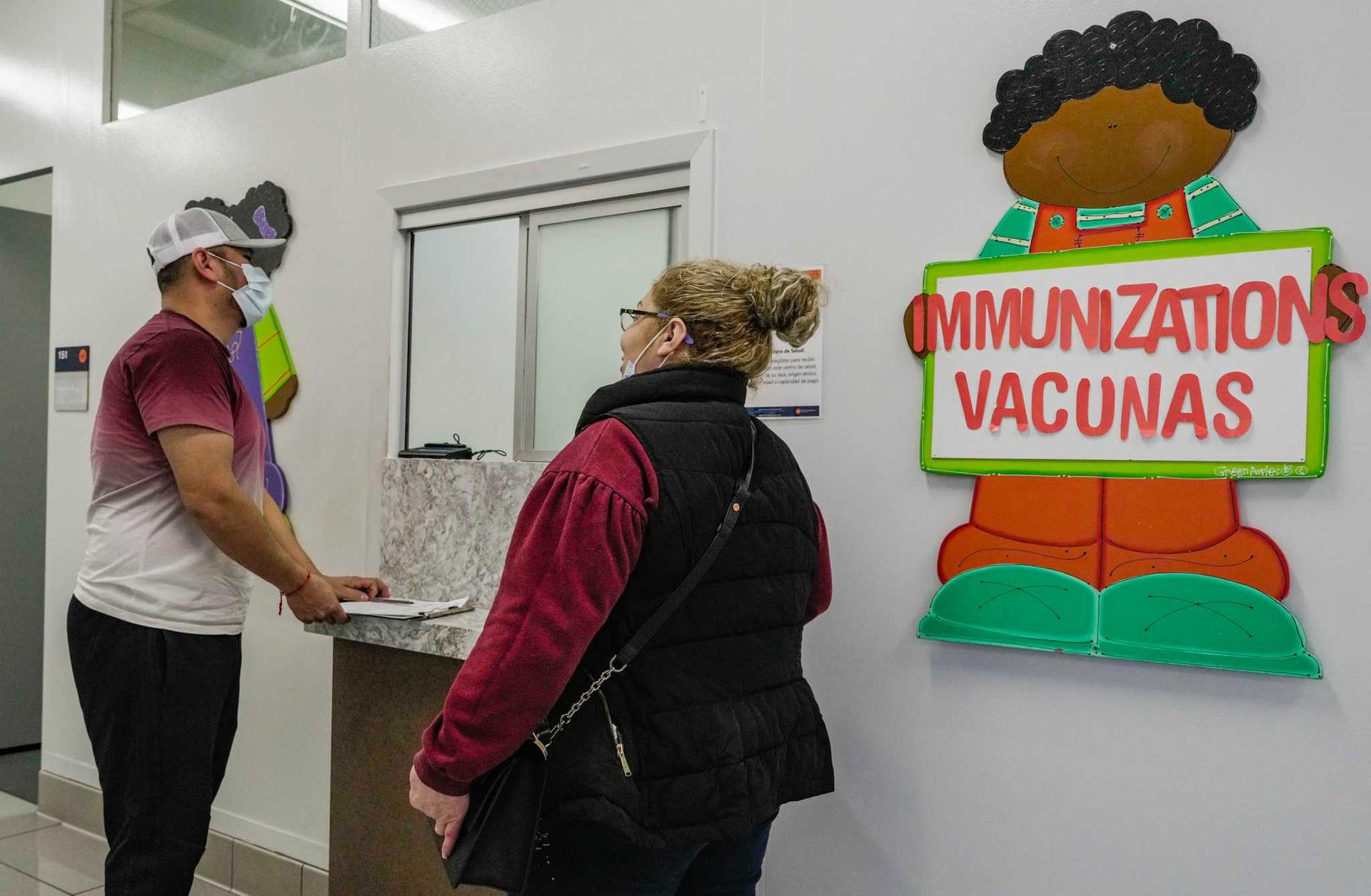 A man and a woman with masks on stand at the front desk of a health clinic. On the wall is a cartoon person holding a sign that reads ''immunizations vacunas.'' 