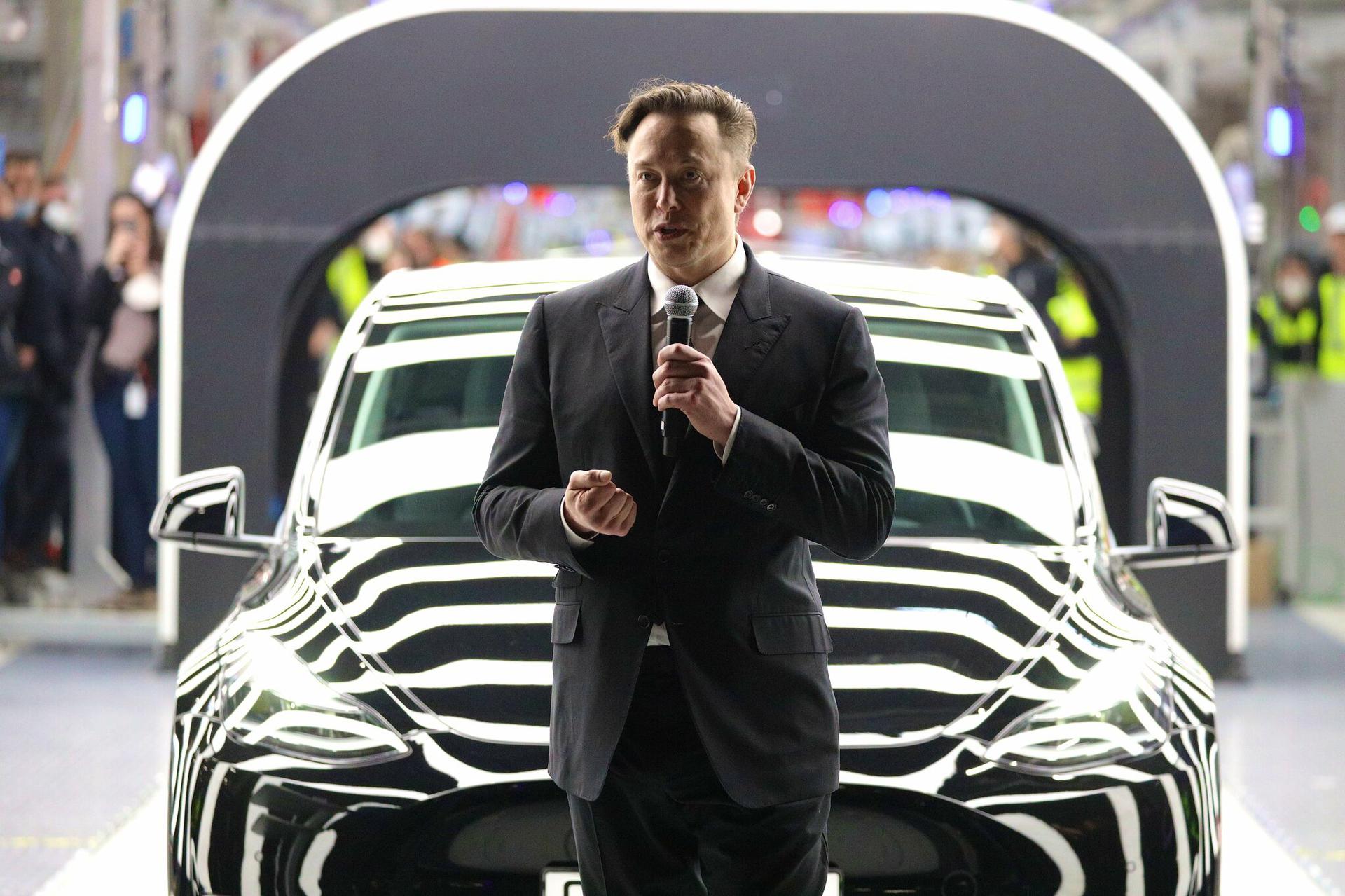 Elon Musk holds a microphone standing in front of a Tesla. 
