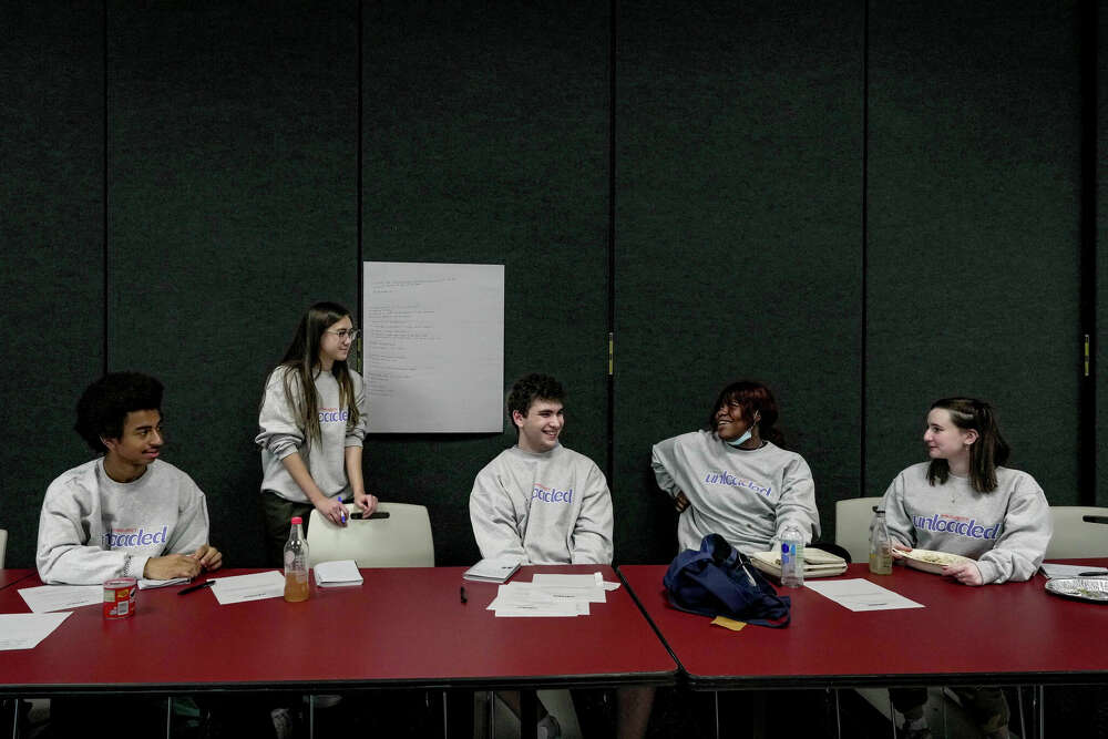 Five young people in matching grey sweaters that say ''project unloaded'' sit at a long table talking. 