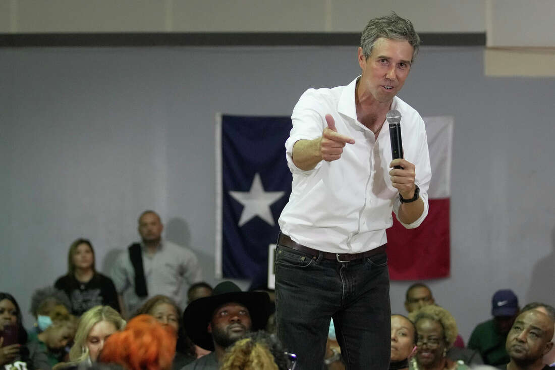 Beto O'Rourke stands with a microphone. Behind him sits a crowd of people. Behind them a Texas flag is hung on the wall. 