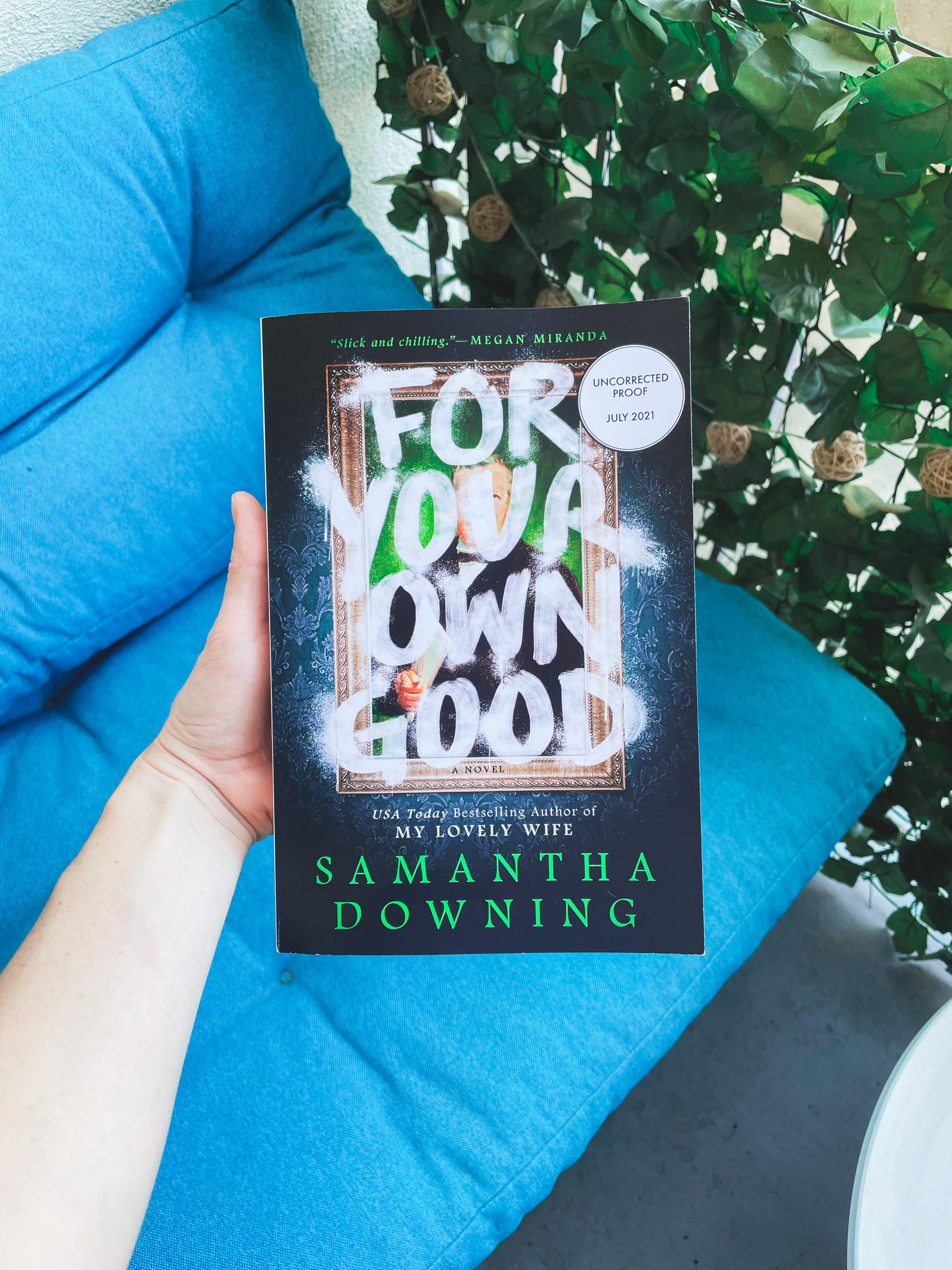A hand holds a copy of ''for your own good'' over a couch next to some plants