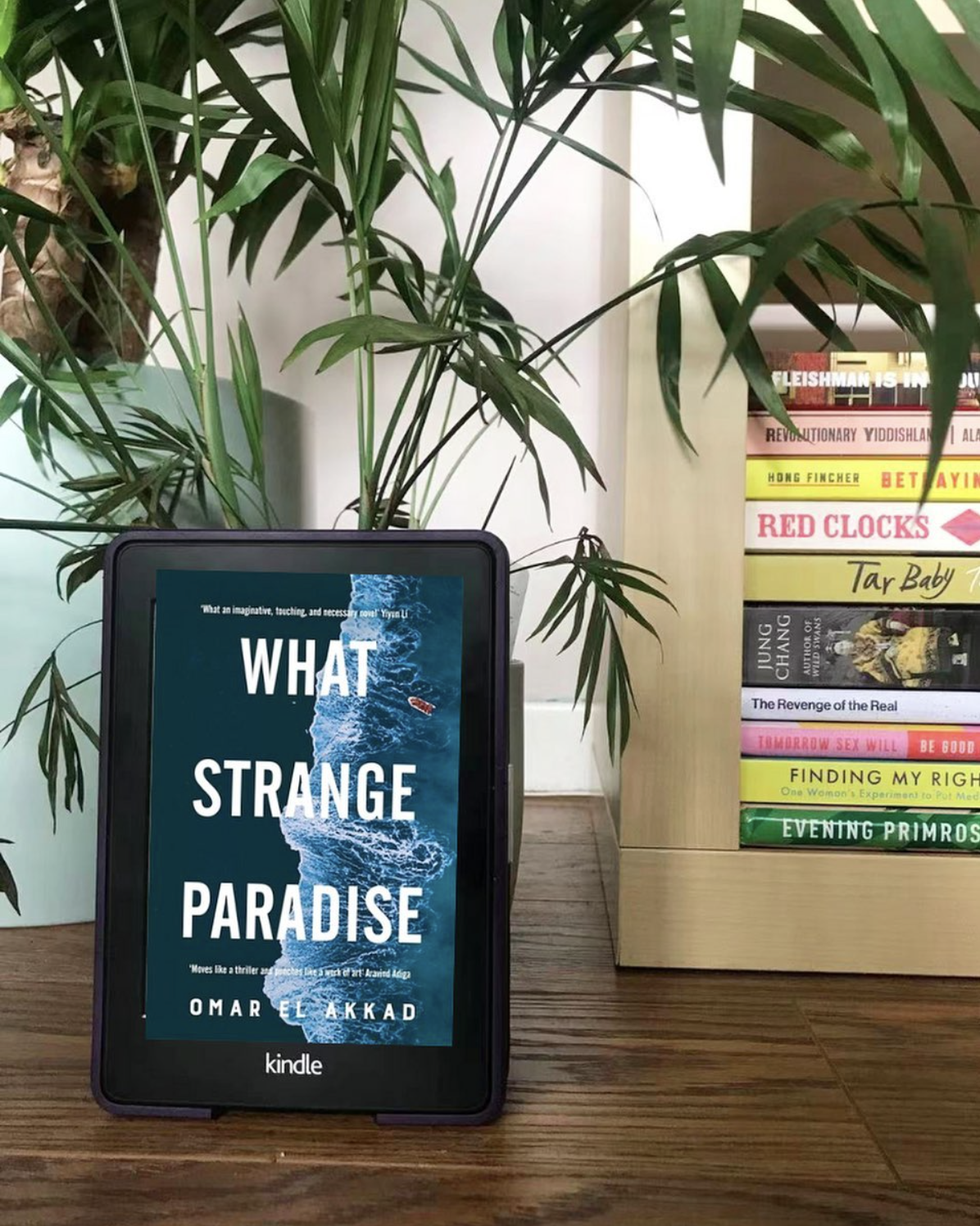 A kindle with the cover of ''what strange paradise'' leans against a plant next to a stack of books A kindle with the cover of ''what strange paradise'' leans against a plant next to a stack of books