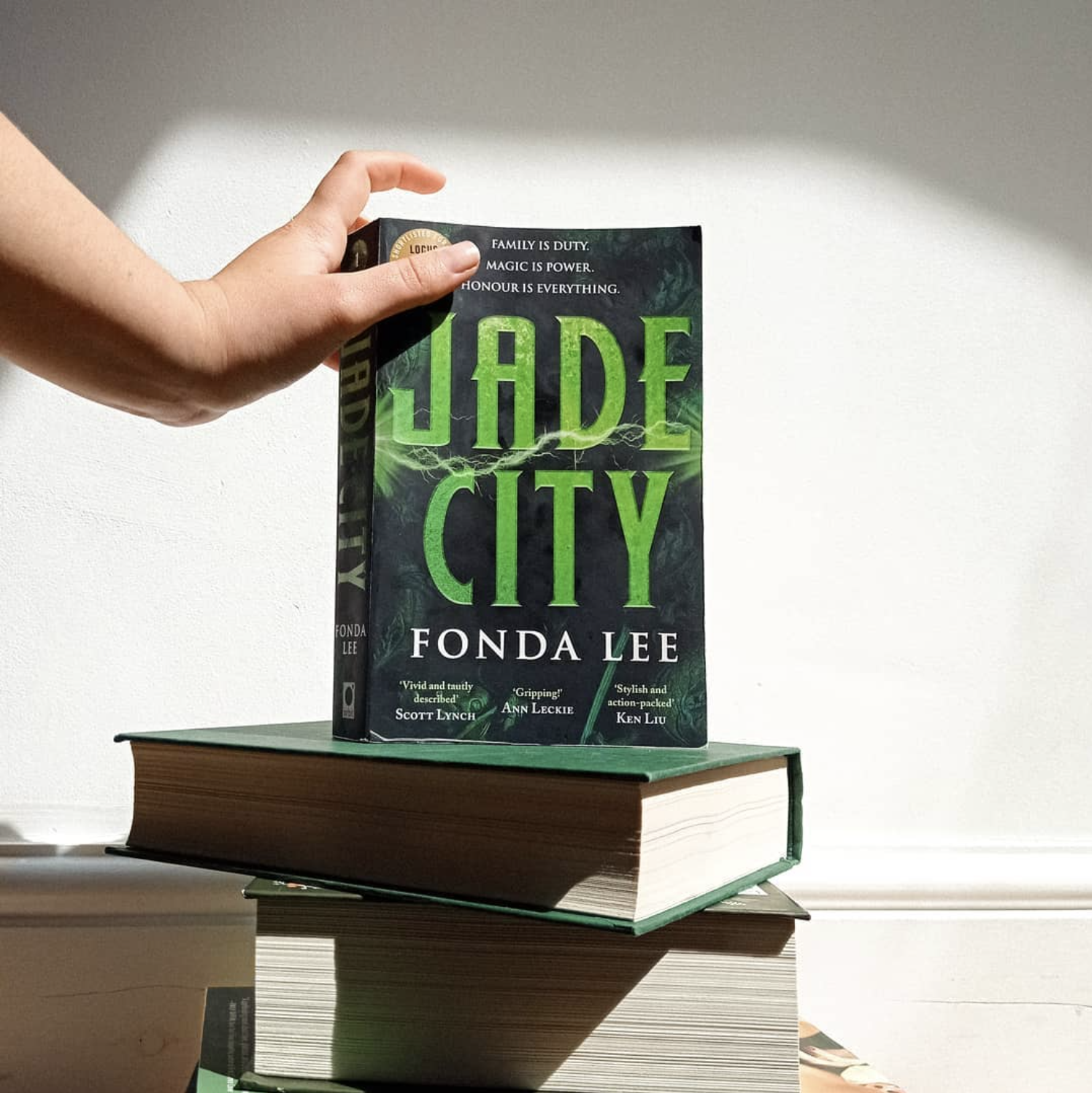A hand holds a copy of ''jade city'' resting on top of a stack of other books A hand holds a copy of ''jade city'' resting on top of a stack of other books