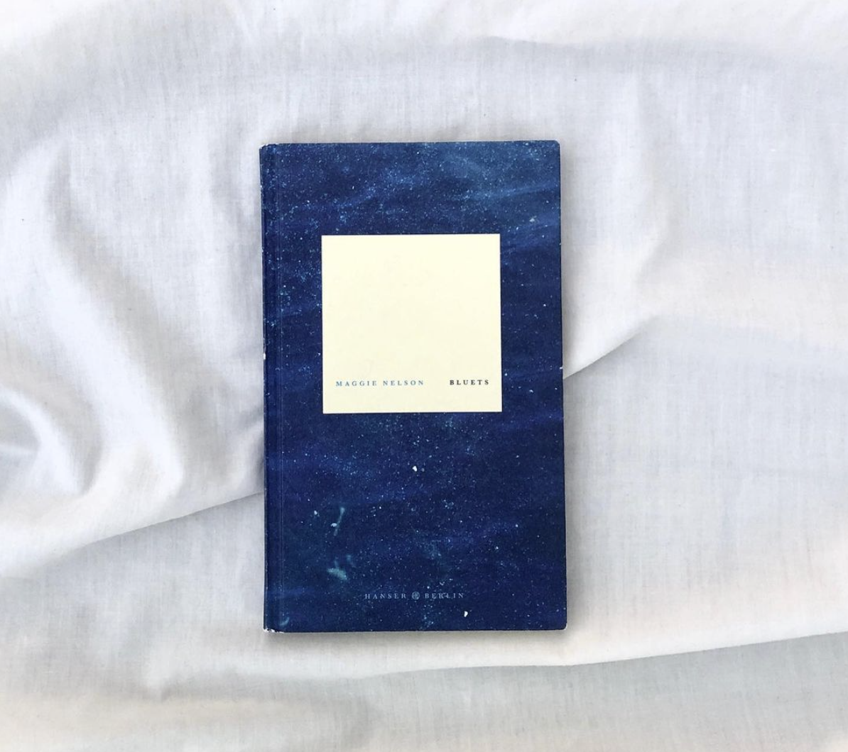 A paperback copy of Bluets flat against a slightly wrinkled linen sheet
