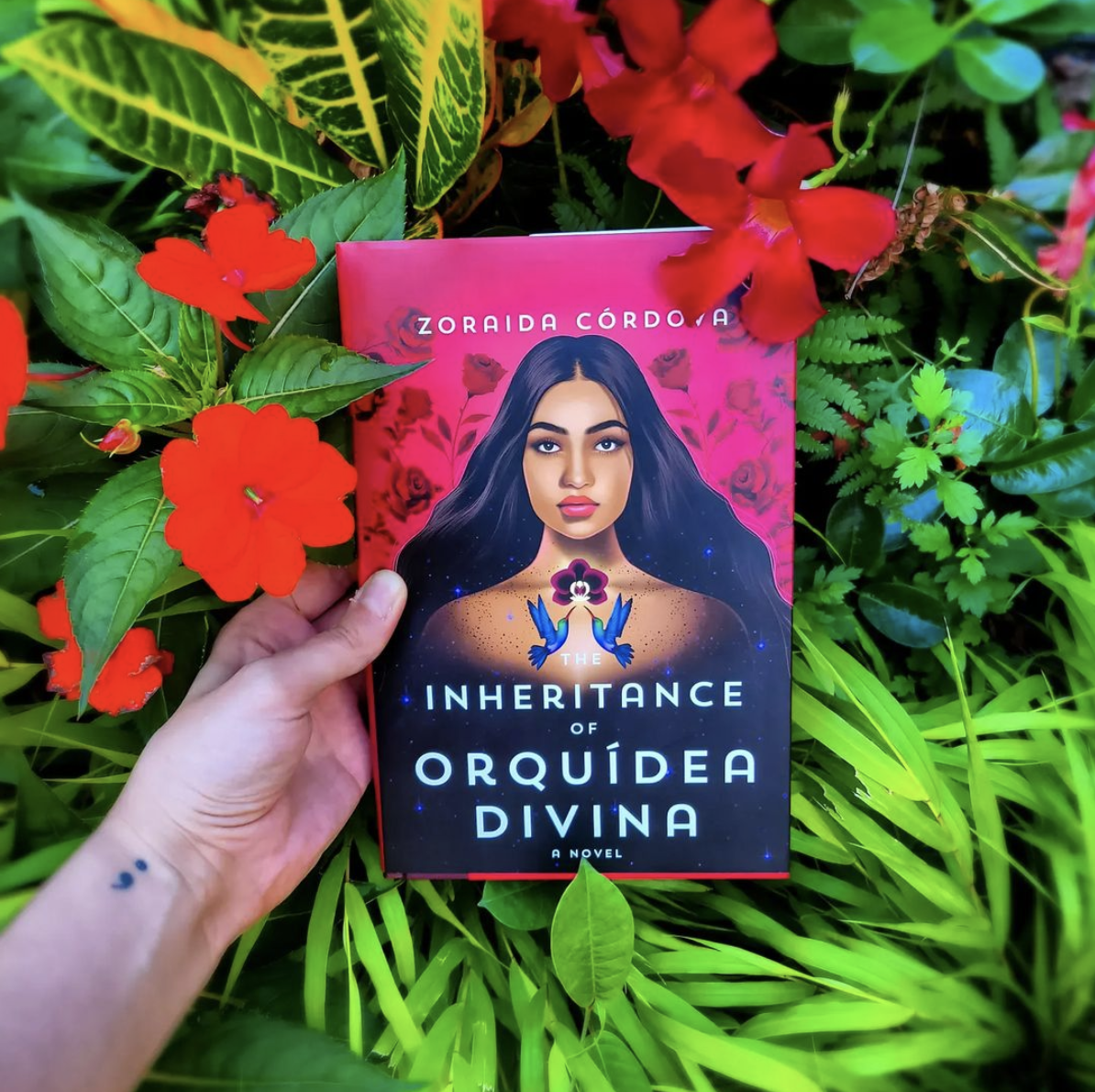 a hand holds a copy of ''the inheritance of orquîdea divina'' above a bed of vibrant flowers and plants a hand holds a copy of ''the inheritance of orquîdea divina'' above a bed of vibrant flowers and plants