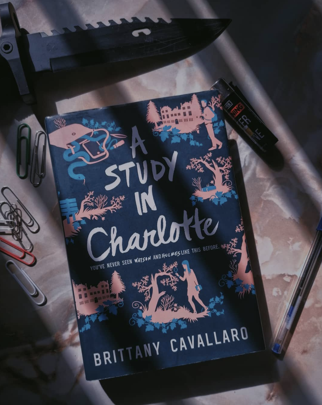 a copy of the book sits in partial shadow surrounded by paper clips, a pocket knife, and a lighter a copy of the book sits in partial shadow surrounded by paper clips, a pocket knife, and a lighter