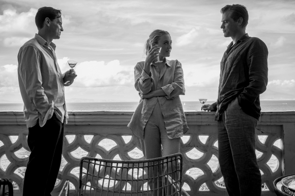 Three people speaking on a balcony overlooking the ocean, each holding a martini.