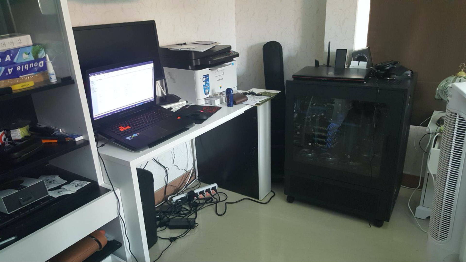 A desk with an open laptop and other computer peripherals, and a large gaming machine next to it
