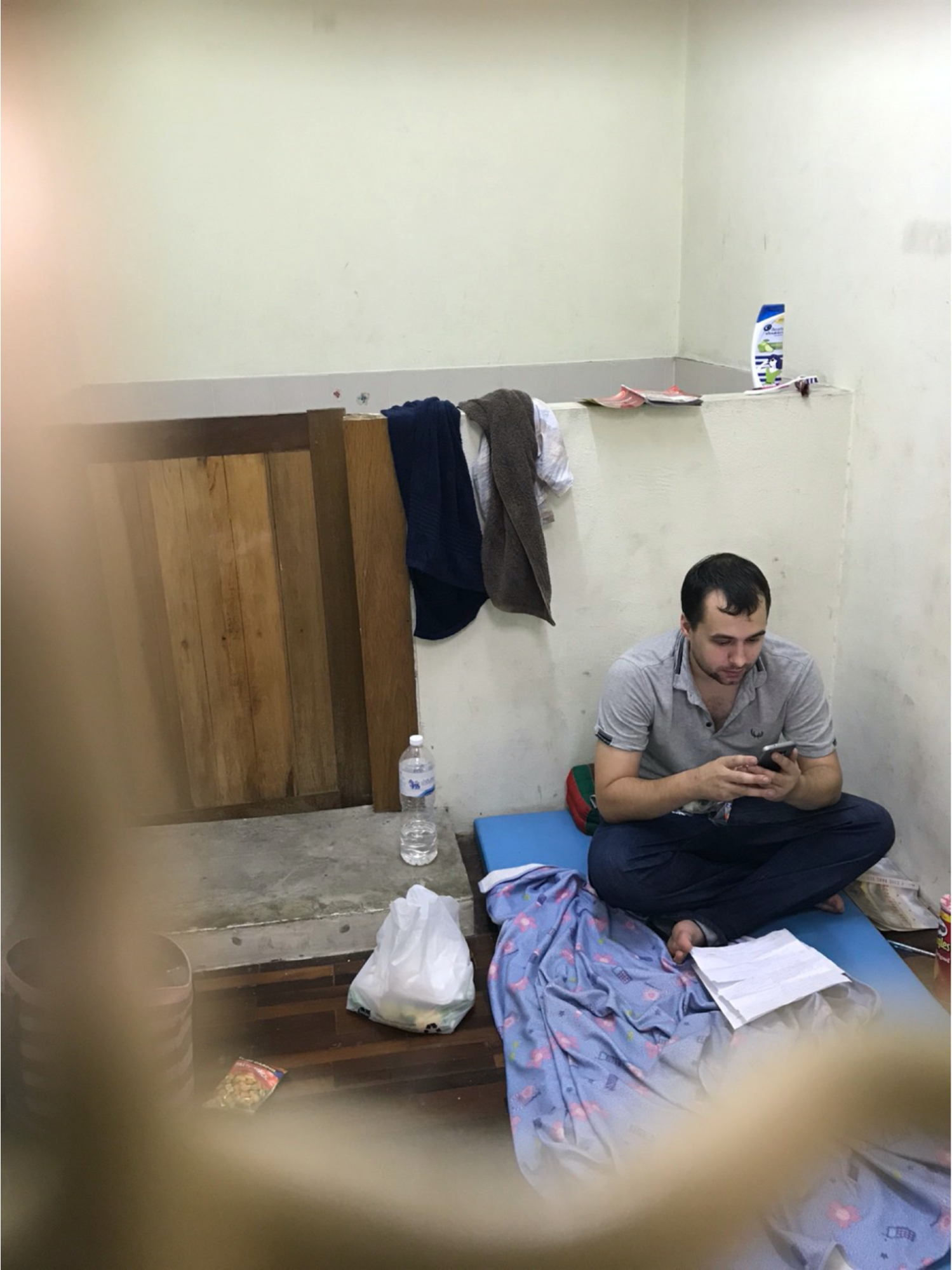 Cazes sitting on a thin mat on the floor and looking at his phone, next to a concrete slab with a water bottle