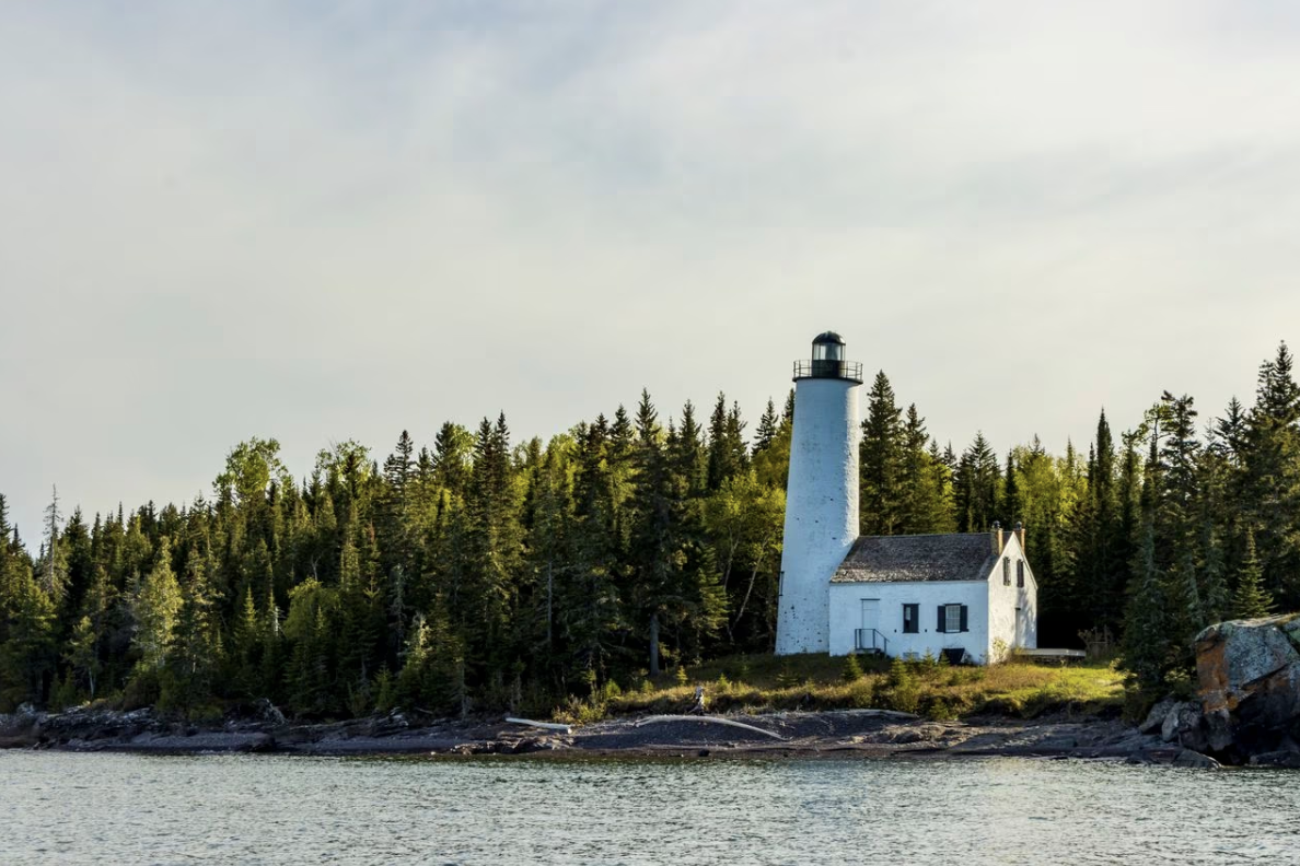 A lighthouse sits on the edge of Lake Superior surrounded by confiers