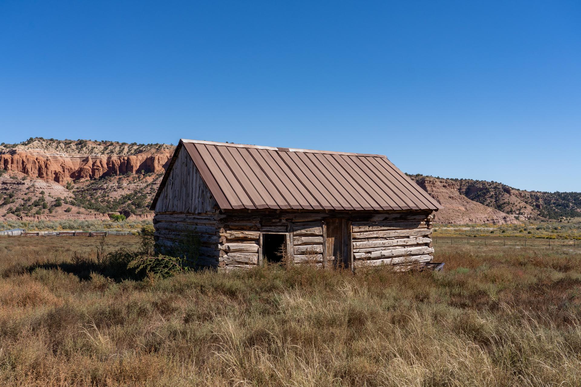 A log cabin in a field, backed by a low ridge.