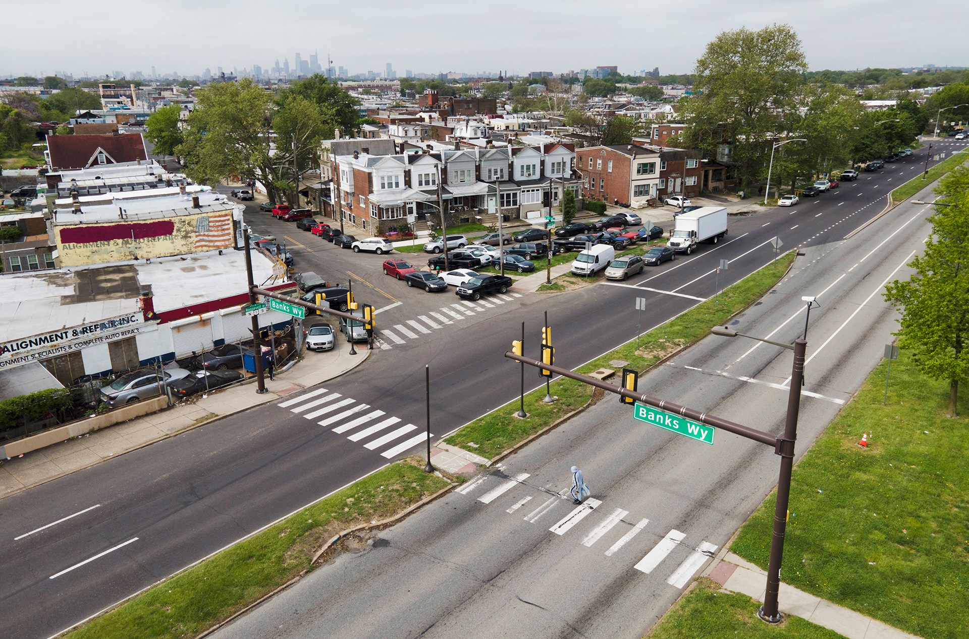 An aerial photo of two multi-lane streets divided by a grassy median. On one side, a town of row-houses and a city skyline are visible. 