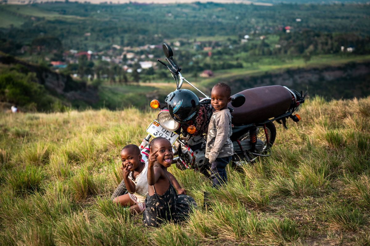 Three children sit with a motorcycle atop a grassy hill with a town in the valley behind them.
