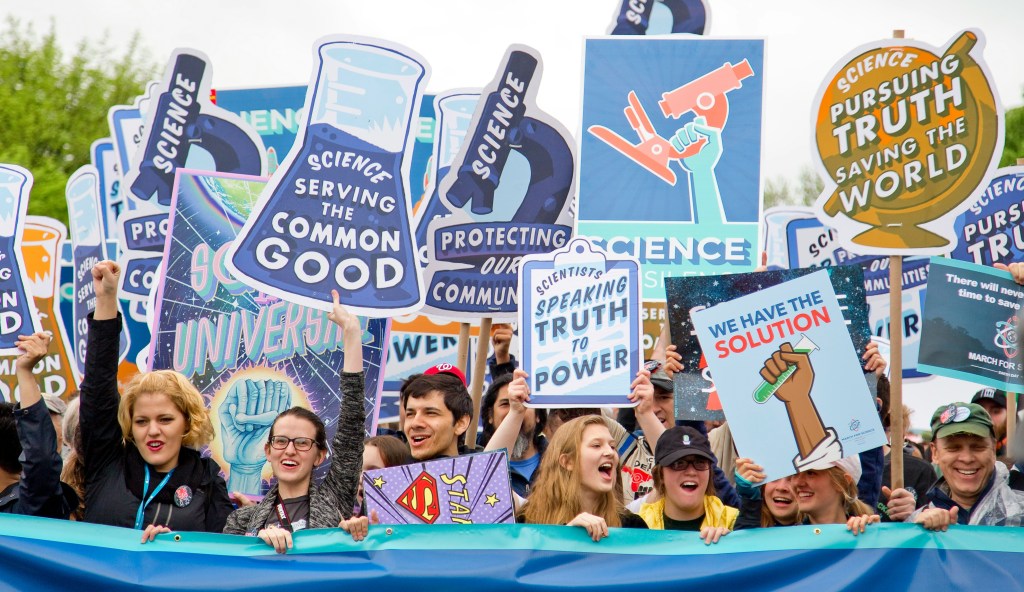 A crowd of demonstrators cheer and hold up blue signs with slogans like: ''Science Serving the Common Good'' and ''Scientists Speaking Truth to Power''