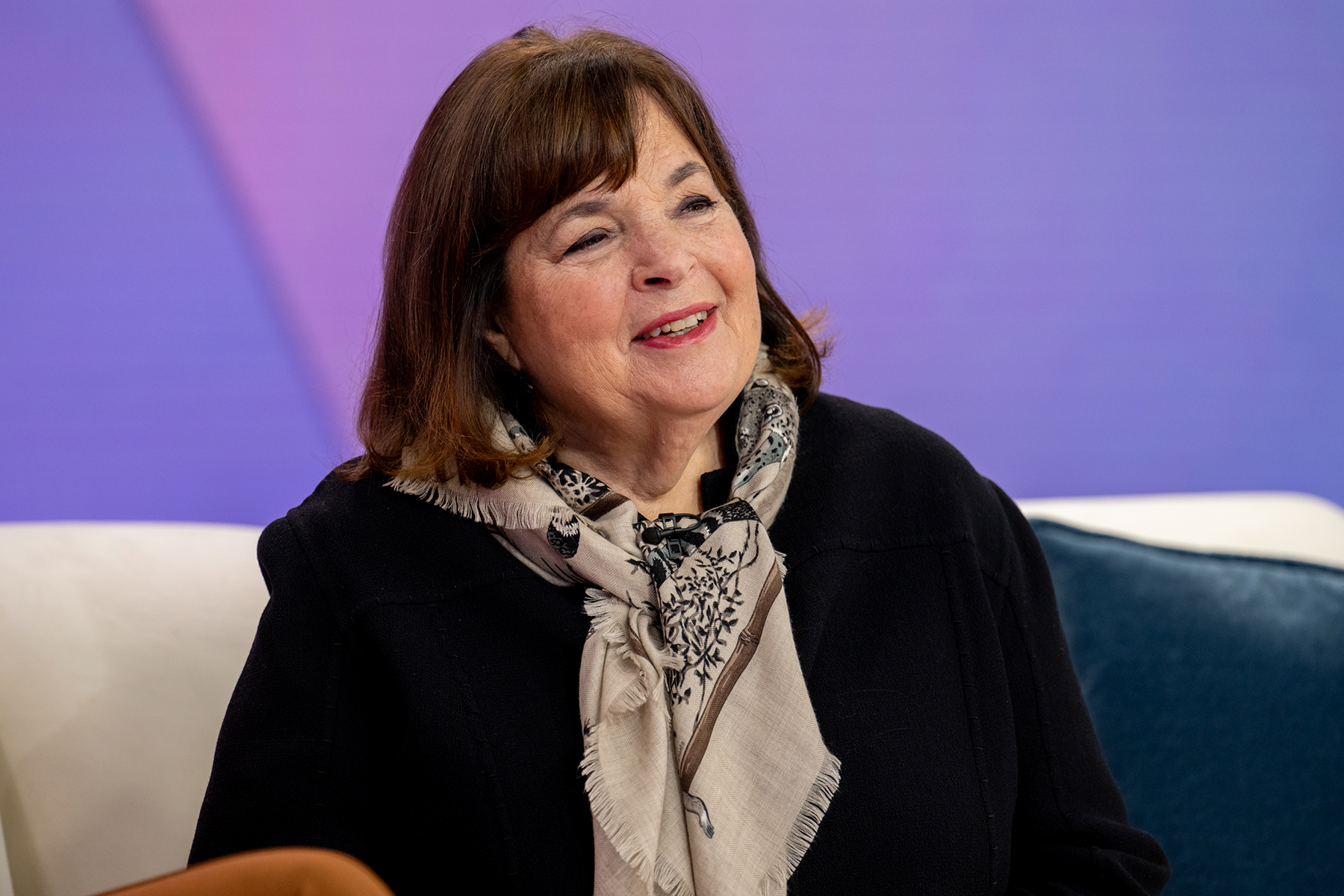 Chef Ina Garten sitting on a couch wearing her signature scarf in tan and black, smiling at someone off screen