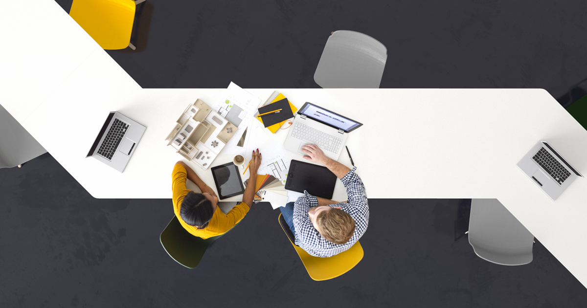 Overhead view of two people collaborating at a white conference table with laptops, documents, and design materials.