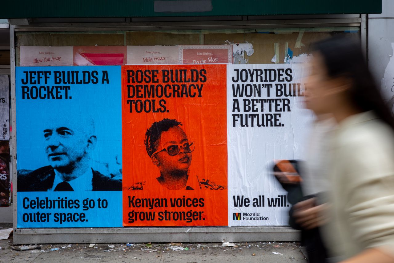 Three posters are pasted side by side on a city wall, and a blurred pedestrian walks past on the right. The blue poster on the left reads "Jeff builds a rocket. Celebrities go to outer space." above a black-and-white photo of Jeff Bezos. The orange poster in the middle reads "Rose builds democracy tools. Kenyan voices grow stronger." above a black-and-white photo of Rose Njeri Tunguru. The white poster on the right reads "Joyrides won't build a better future. We all will." and has the Mozilla Foundation logo in the bottom corner.