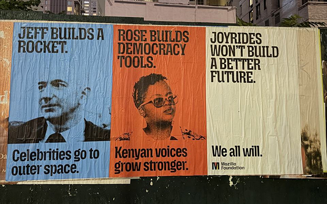 Three posters pasted side by side on a dark green construction wall at night, with lit New York high-rises behind them. The blue poster on the left reads “Jeff builds a rocket. Celebrities go to outer space.” above a black-and-white photo of Jeff Bezos. The orange poster in the middle reads “Rose builds democracy tools. Kenyan voices grow stronger.” above a black-and-white photo of Rose Njeri Tunguru in glasses. The white poster on the right reads “Joyrides won’t build a better future. We all will.” and has the Mozilla Foundation logo in the bottom corner.