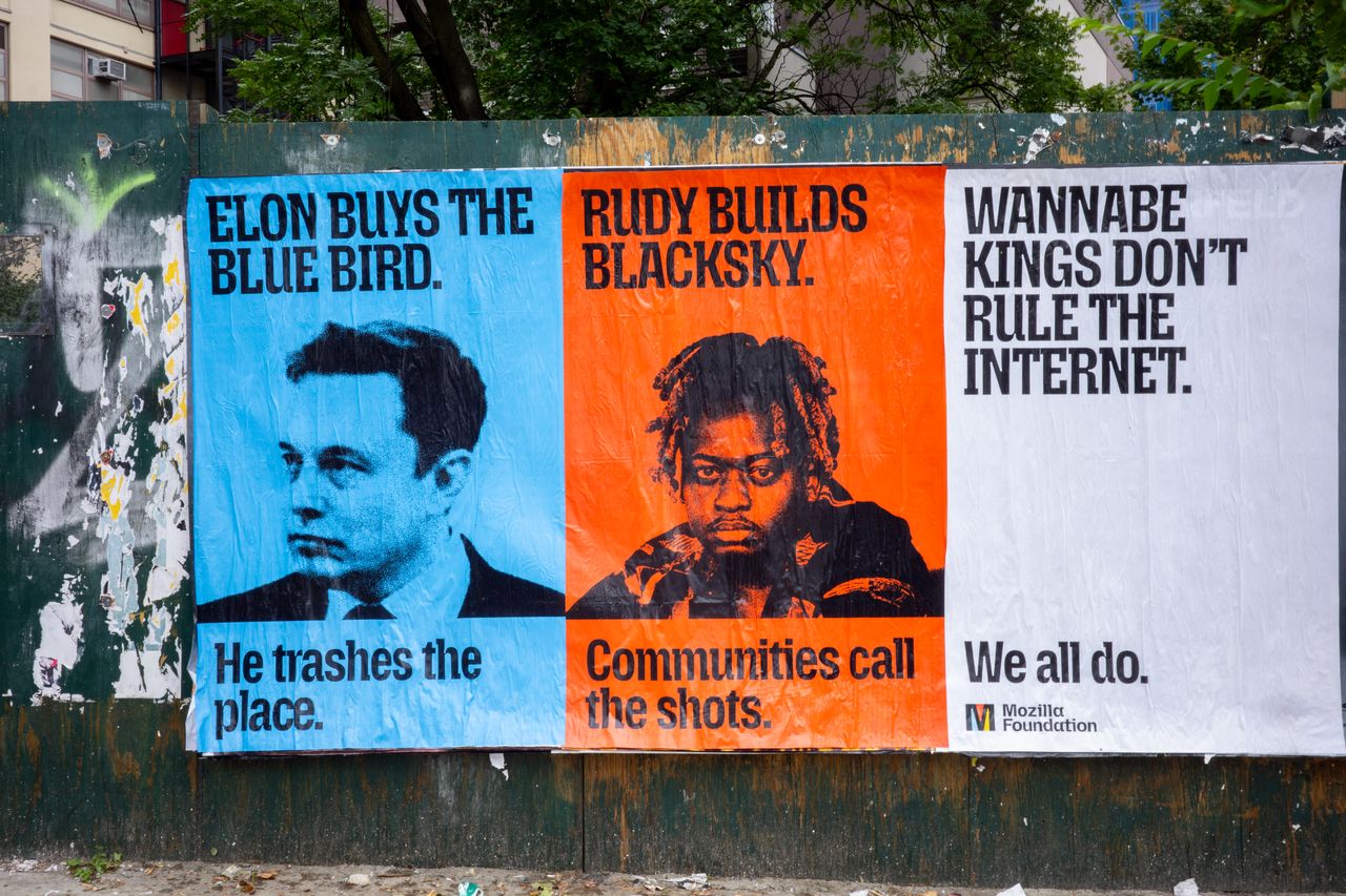 Three posters are pasted side by side on a weathered green construction wall with trees and buildings behind it. The blue poster on the left reads "Elon buys the blue bird. He trashes the place." above a black-and-white photo of Elon Musk. The orange poster in the middle reads "Rudy builds BlackSky. Communities call the shots." above a black-and-white photo of Rudy Fraser. The white poster on the right reads "Wannabe kings don't rule the internet. We all do." and has the Mozilla Foundation logo in the bottom corner.