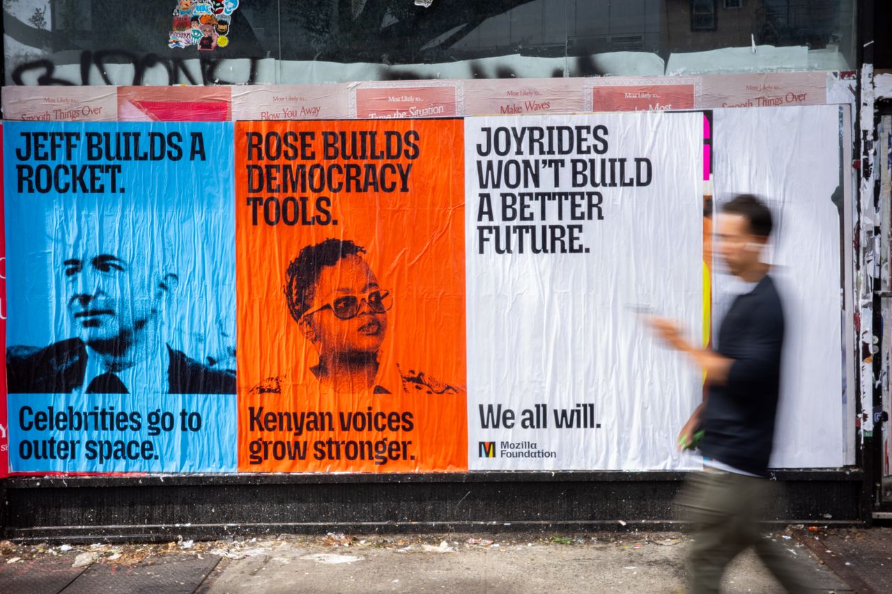 Three posters are pasted side by side on a city wall, and a blurred pedestrian walks past on the right. The blue poster on the left reads "Jeff builds a rocket. Celebrities go to outer space." above a black-and-white photo of Jeff Bezos. The orange poster in the middle reads "Rose builds democracy tools. Kenyan voices grow stronger." above a black-and-white photo of Rose Njeri Tunguru. The white poster on the right reads "Joyrides won't build a better future. We all will." and has the Mozilla Foundation logo in the bottom corner.