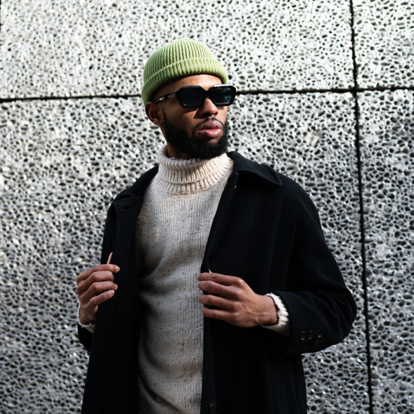 man wearing a dress coat over a sweater, with sunglasses and a green hat