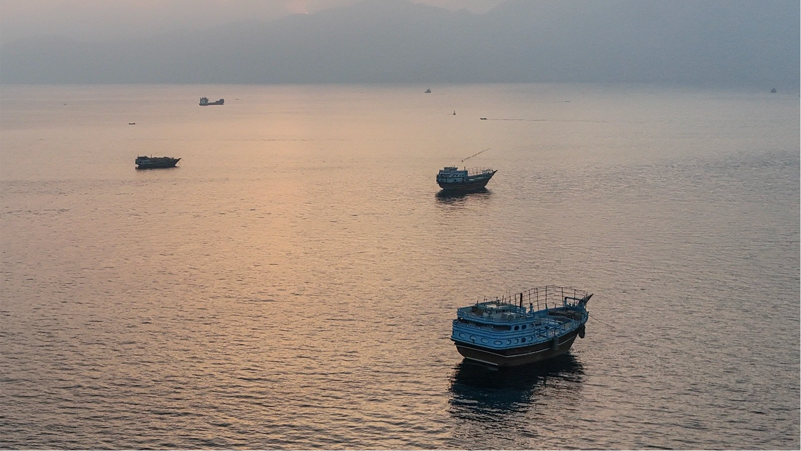 Boats sit in the water with the sun low in a cloudy sky.