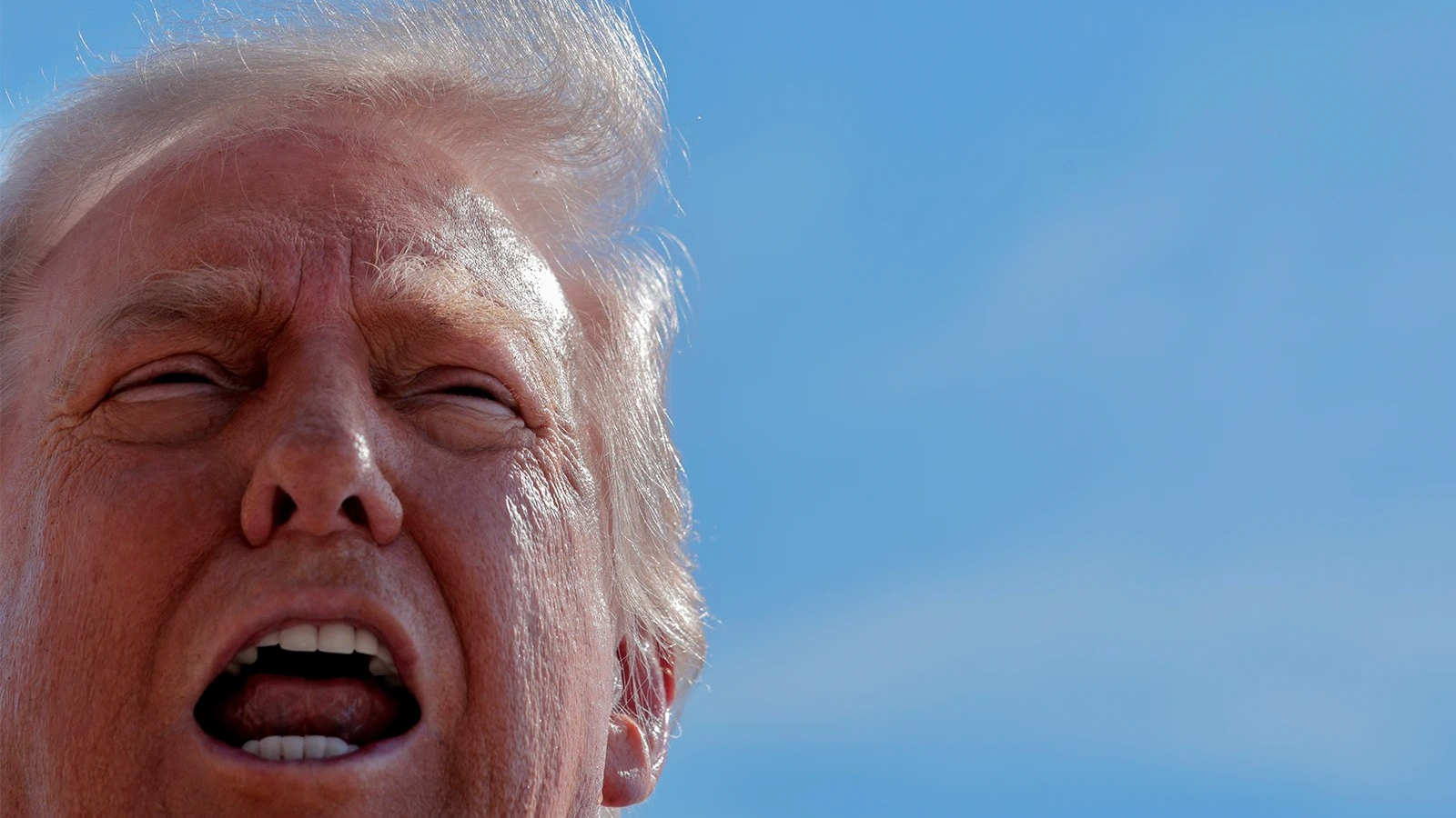 Donald Trump — just his face visible — with his mouth open. He is outside and seen against the blue sky.