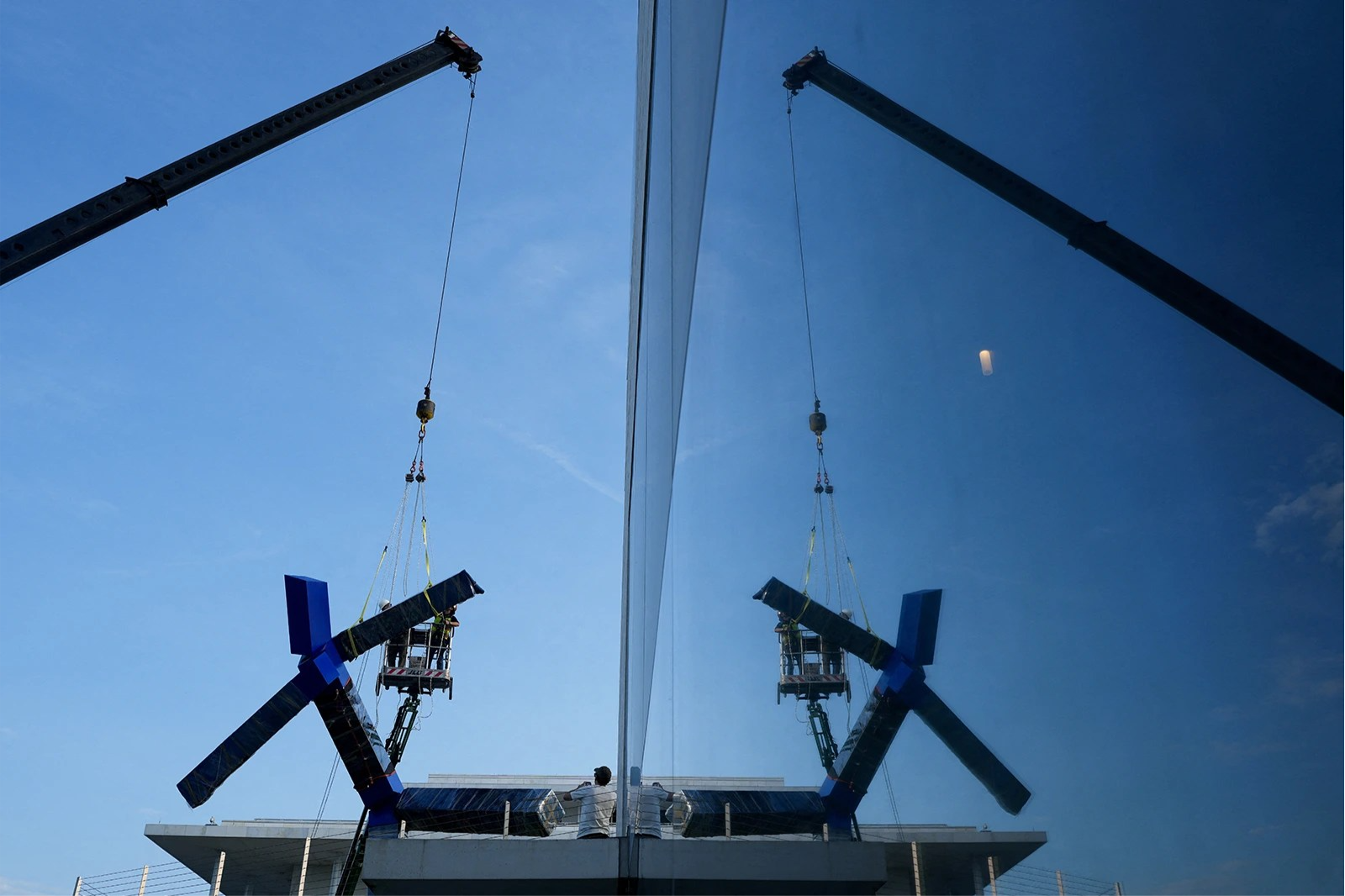 Workers with a crane take apart a large blue statue. The scene is reflected in a nearby window.
