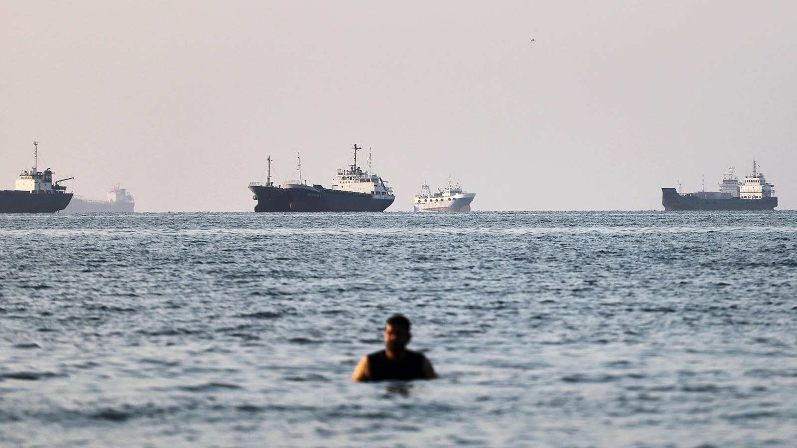 A man stands in the sea while five ships can be seen in the background