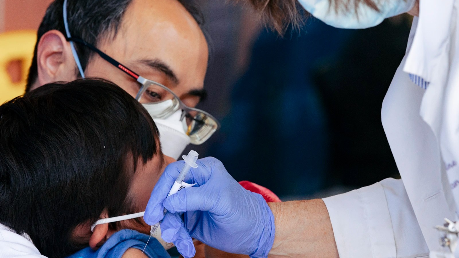 A kid, left, recoils in the arms of his father, center. A nurse, right, sticks a needle in the shoulder of the kid. No faces are completely visible.