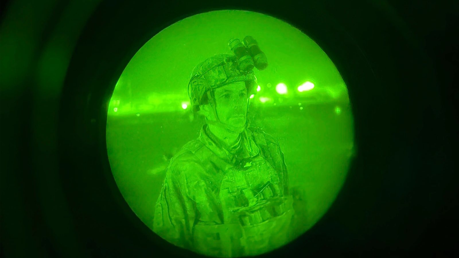 A person wearing army gear is seen through a night vision scope, which looks like him in a green circle surrounded by all black.