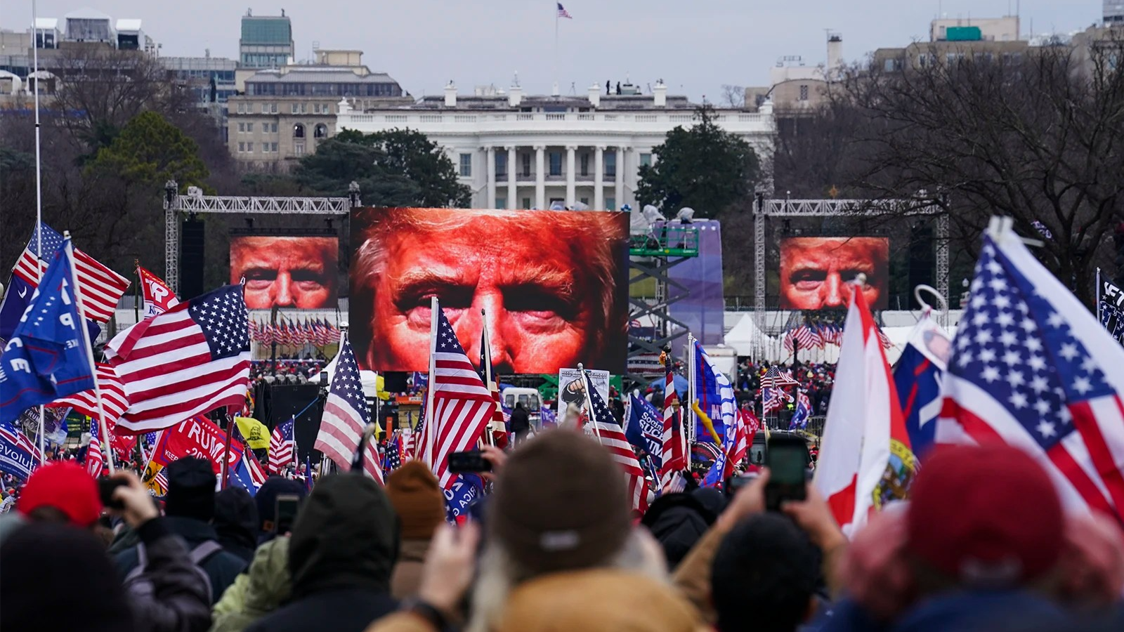 Donald Trump's face is seen on multiple big screens as his supporters stand in the foreground holding flags.