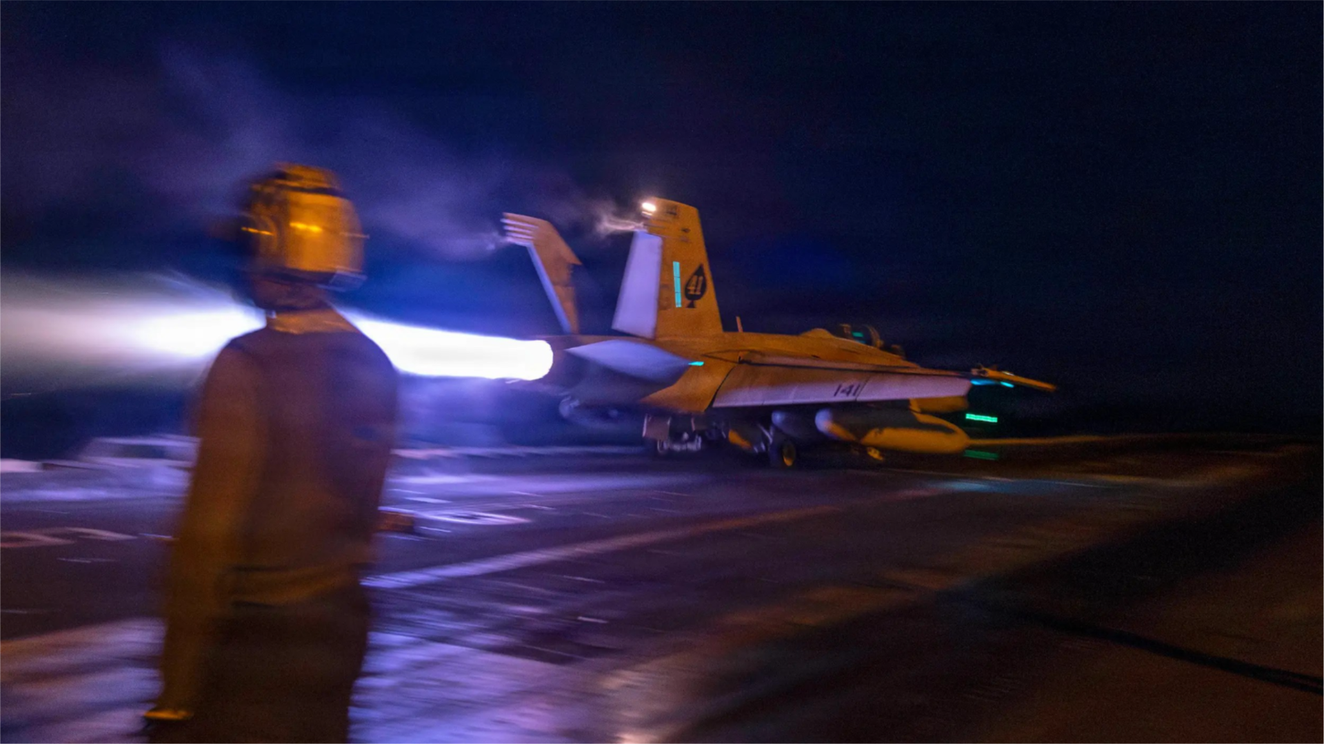 A fighter jet's engines shoot fire as a crew member watches